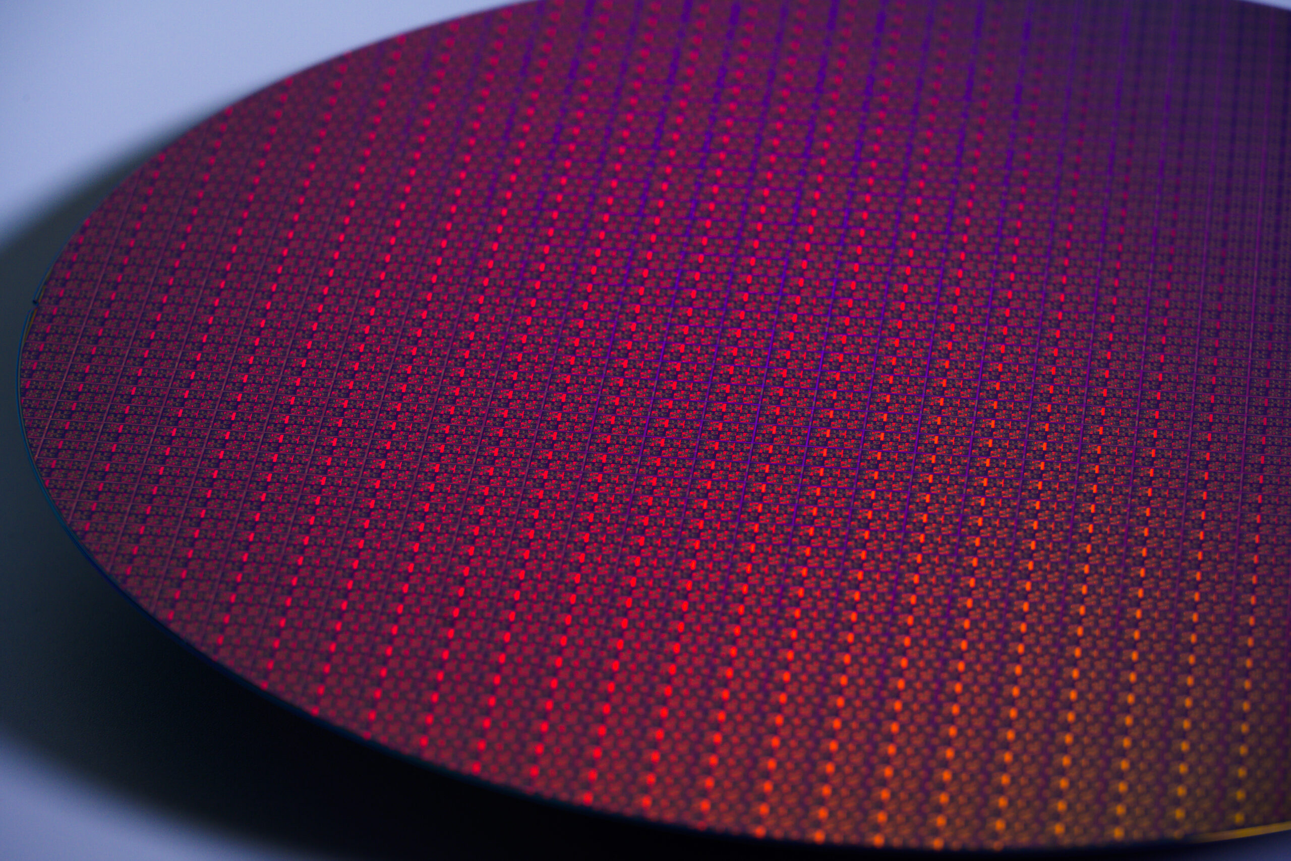 Close-up of a vibrant semiconductor wafer, displaying a grid pattern of tiny, multicolored squares. The surface reflects hues of red, purple, and orange, creating a colorful and intricate mosaic effect under the light.