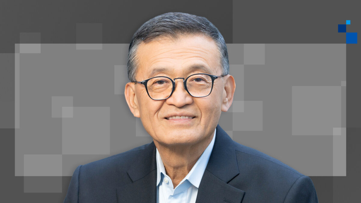 A smiling man with glasses and short hair wearing a dark suit and white shirt, set against a gray background with pixelated squares.